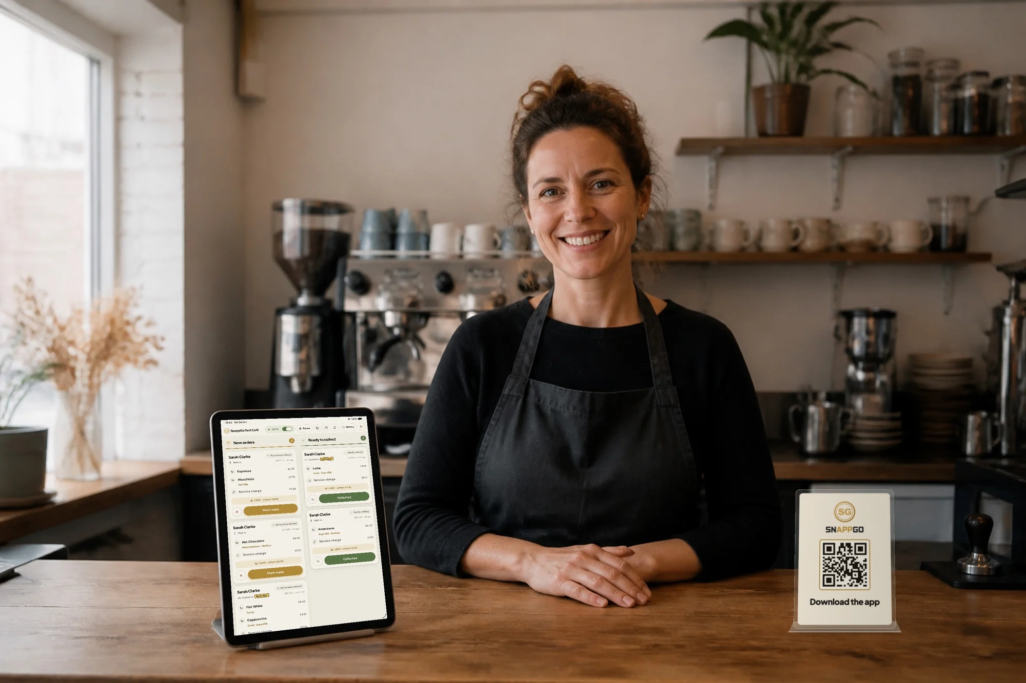 A barista at a café counter next to a tablet showing the SnappGo order board, with a SnappGo Download the app QR code stand on the counter