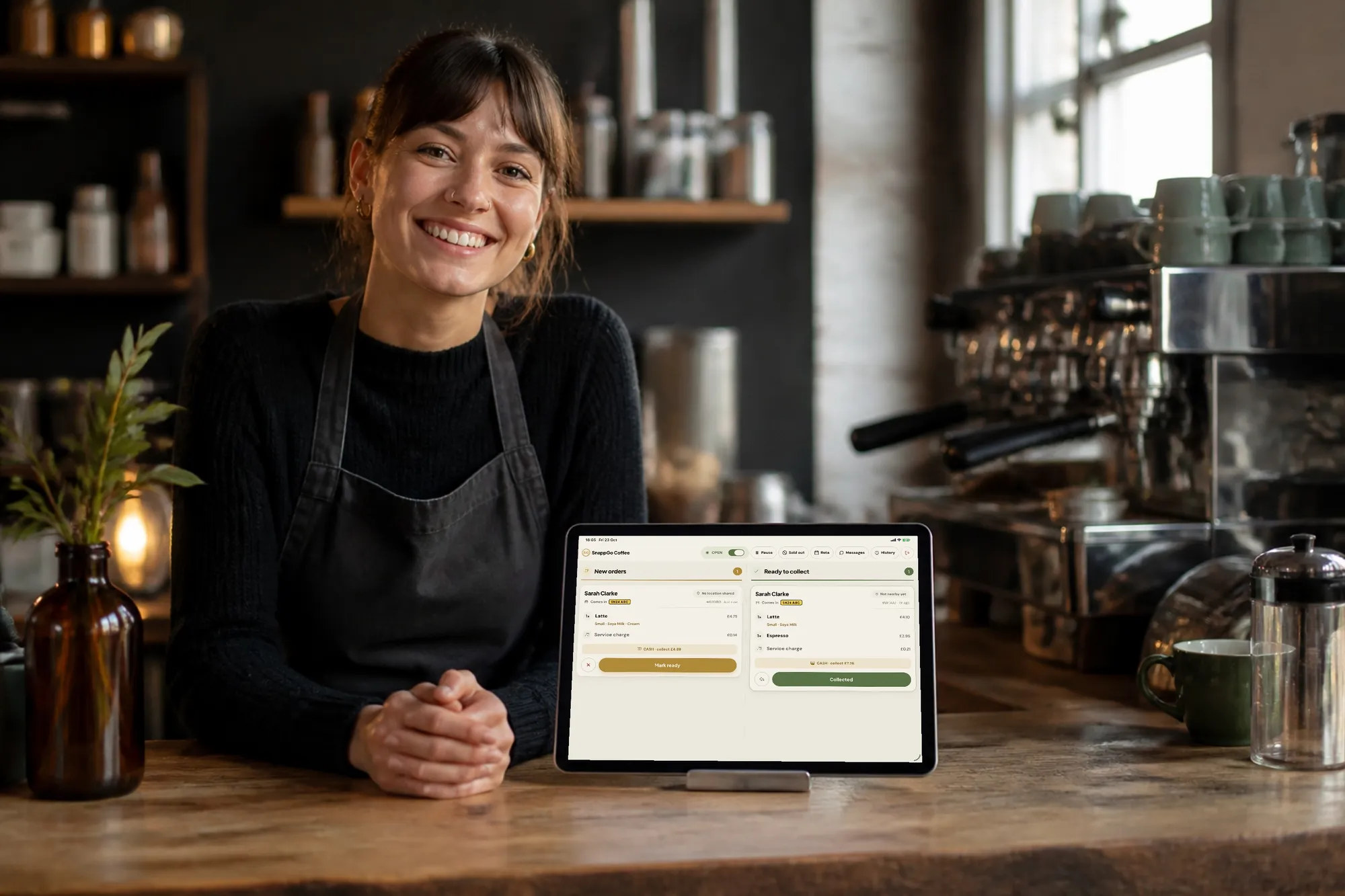 A barista at a café counter next to a tablet showing the SnappGo order board with new orders and orders ready to collect