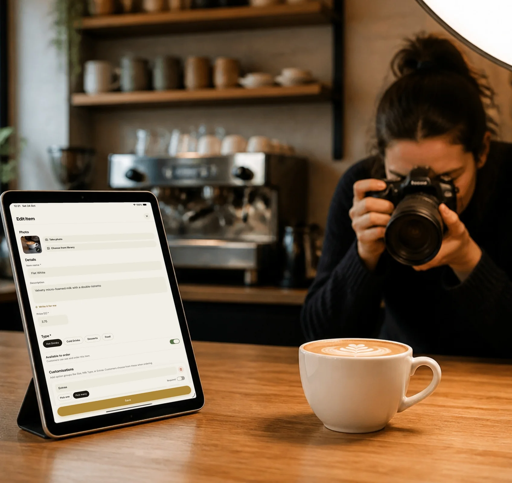 A photographer taking a picture of a latte on a café counter, next to a tablet showing the SnappGo Edit item screen for a Flat White